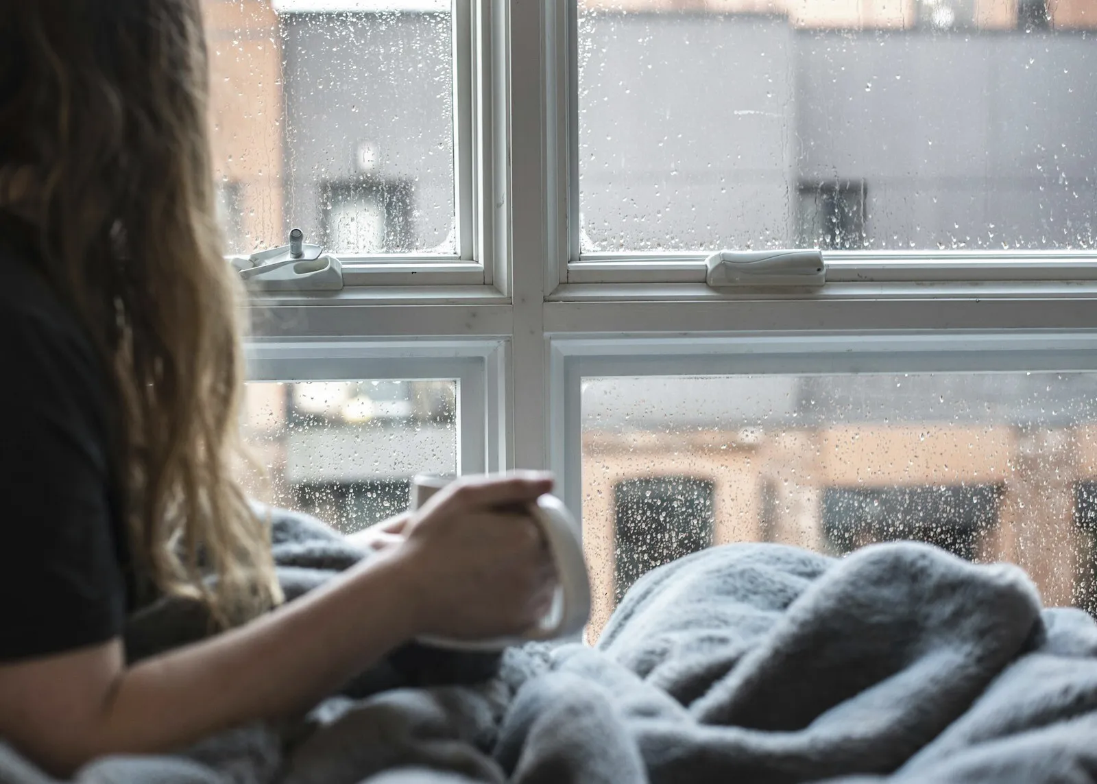 A person sitting by a rain-streaked window holding a mug