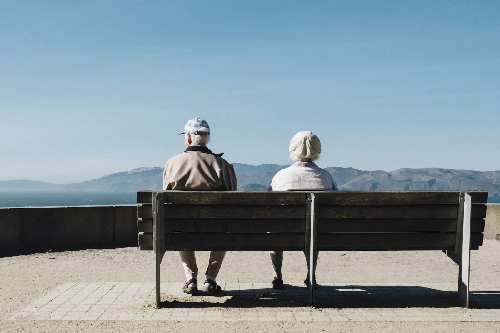 An older couple sitting apart on a bench, looking out